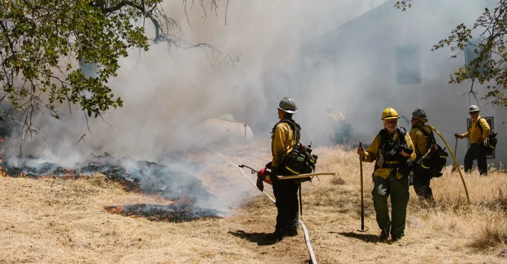 (Firefighters in yellow gear work to contain a brush fire near a home, surrounded by smoke and dry grass—capturing the intensity and urgency of wildfire response efforts.)