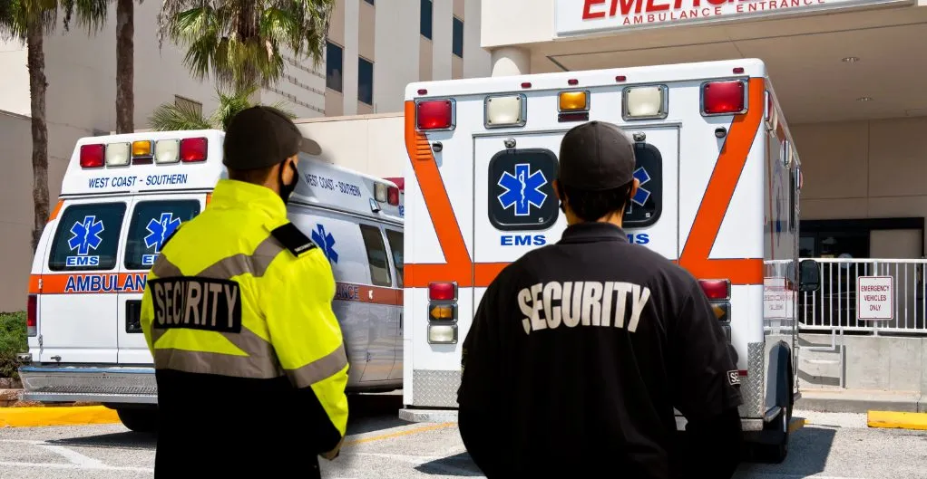 (Two security guards stand watch near ambulances outside a hospital emergency entrance, symbolizing active protection and safety in medical facilities.)