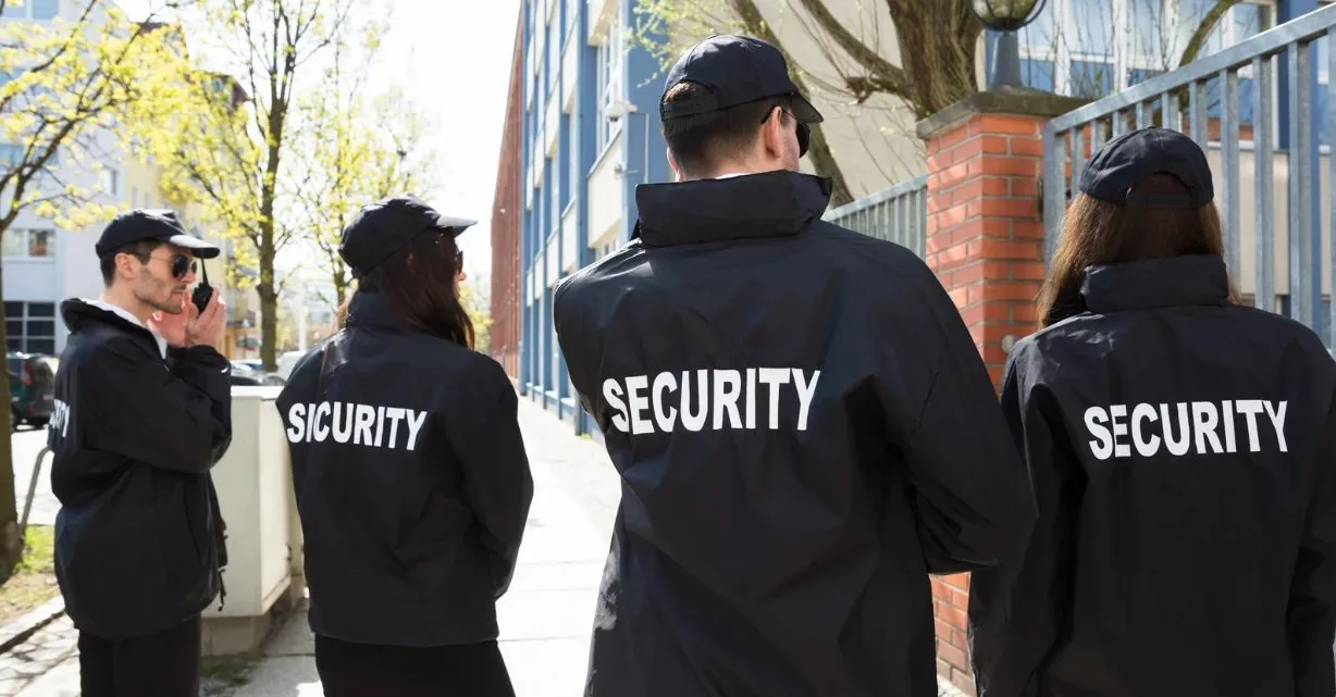 (A group of four security personnel in black jackets conduct an outdoor patrol along a city sidewalk near a gated building.)
