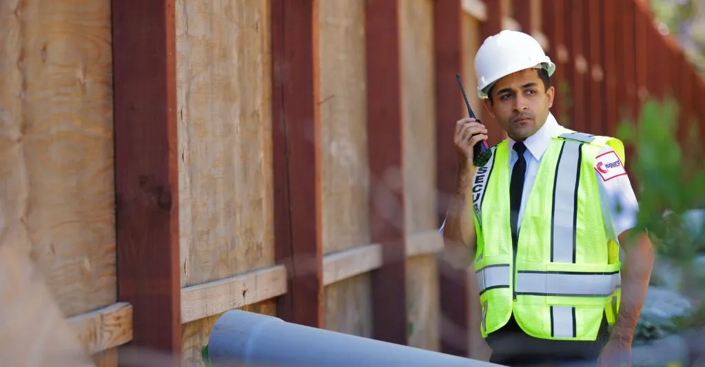 (A security guard in a high-visibility vest and hard hat communicates via radio while patrolling a construction site perimeter, ensuring safety and protection.)