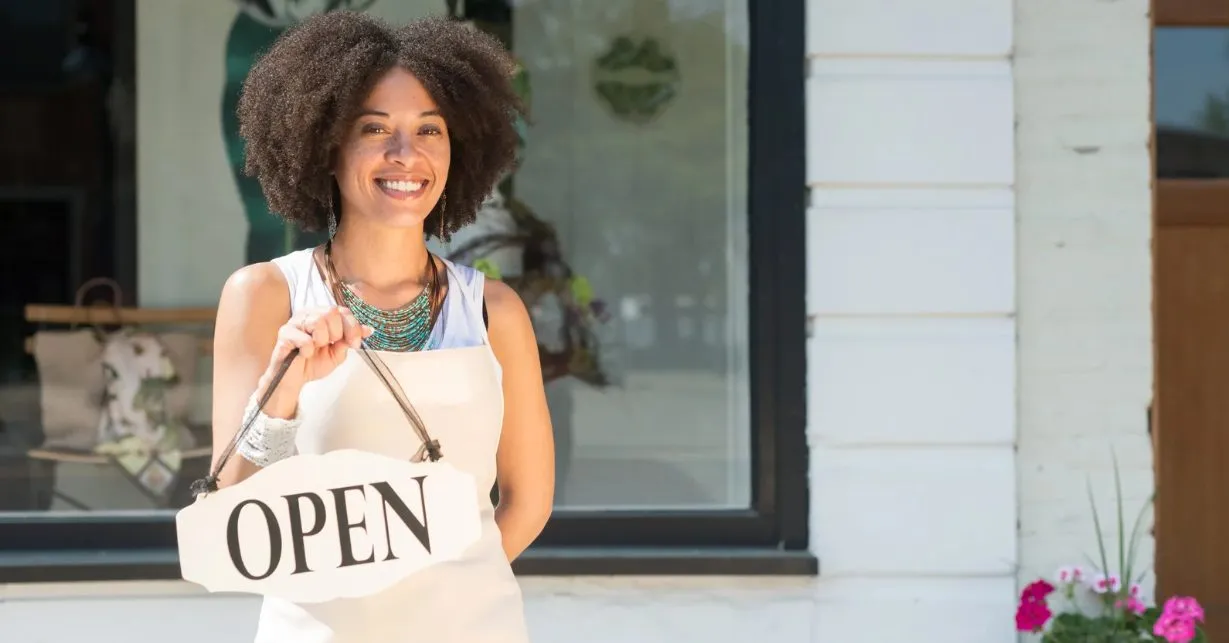 (A smiling shop owner stands outside her storefront holding an “Open” sign, ready to welcome customers.)