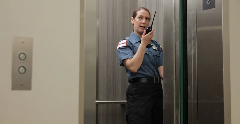 (A female security officer communicates via radio while standing in an elevator, reflecting professional vigilance and responsiveness in facility security operations.)