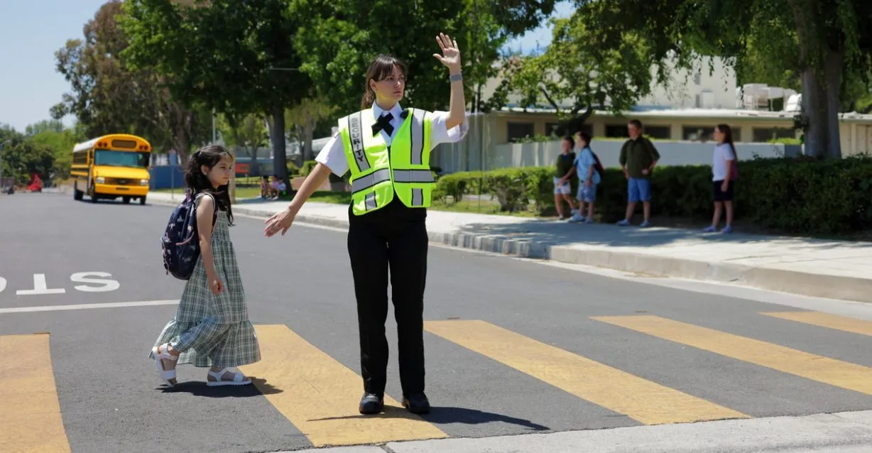 (A Citiguard security officer helps a young student safely cross the street near a school zone, with other children waiting on the sidewalk.)
