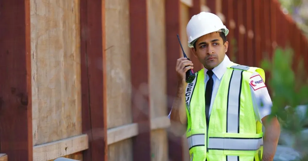 (A construction site security guard in a hard hat and reflective vest communicates via two-way radio while monitoring the perimeter of a job site.)